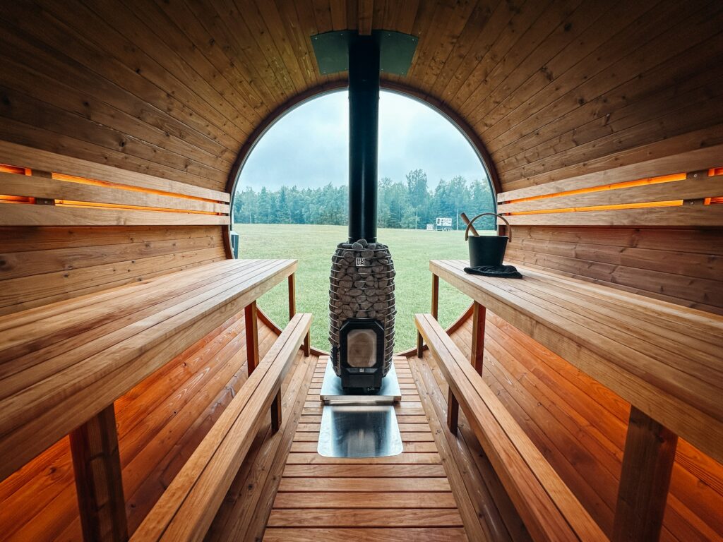 Blick durch eine Fasssauna mit Holzbänken auf den Holzofen vor dem Panoramafenster