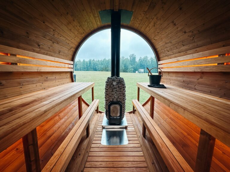 Blick durch eine Fasssauna mit Holzbänken auf den Holzofen vor dem Panoramafenster