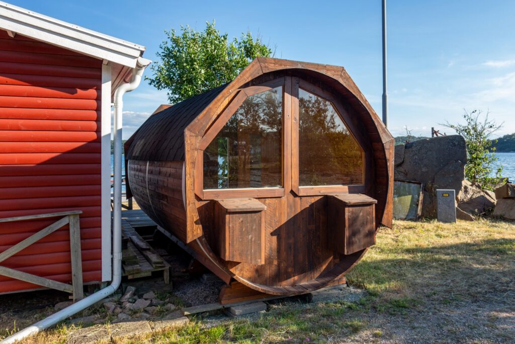 Fasssauna mit großer Glasfront neben einem roten Holzhaus am Meer